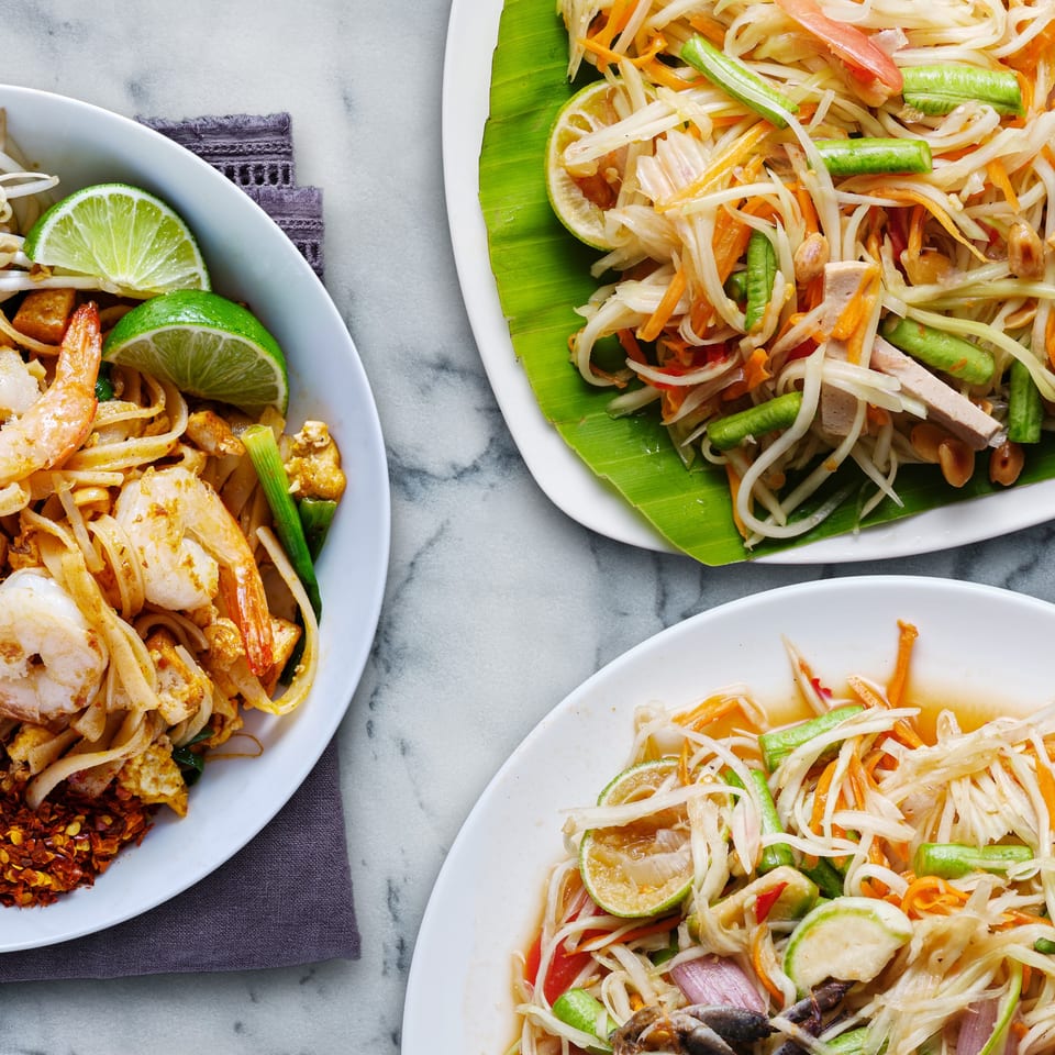 Three plates of Thai food, including shrimp pad Thai and two papaya salads, on a marble surface.
