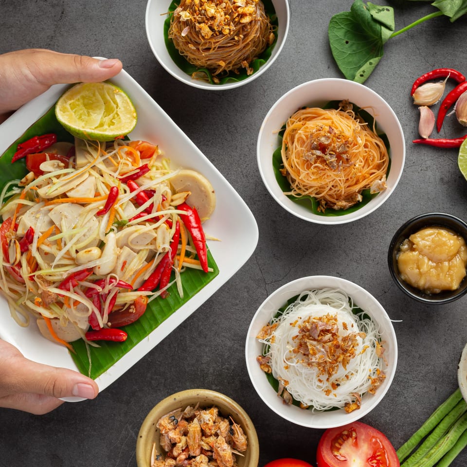 Hands holding a plate of Thai papaya salad with noodles, chili, and lime on a dark background.