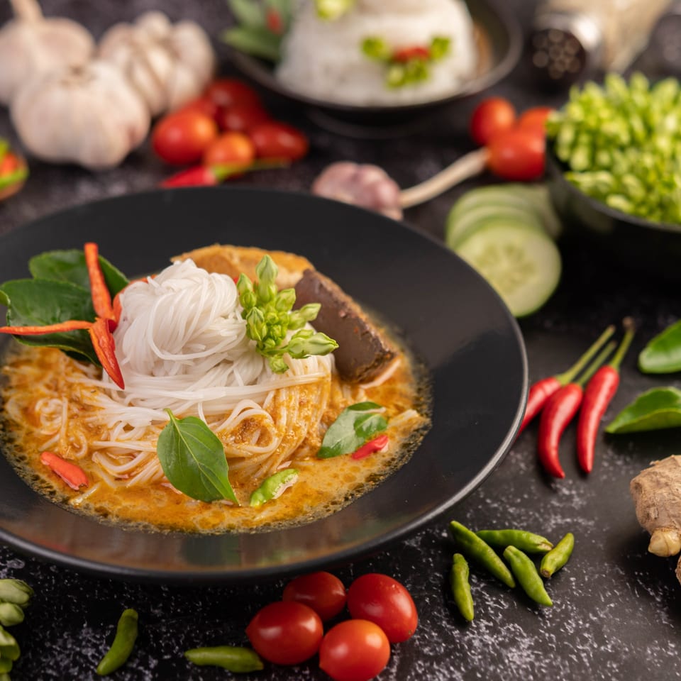 Thai red curry with rice noodles, garnished with herbs, chilies, and vegetables on a dark surface.
