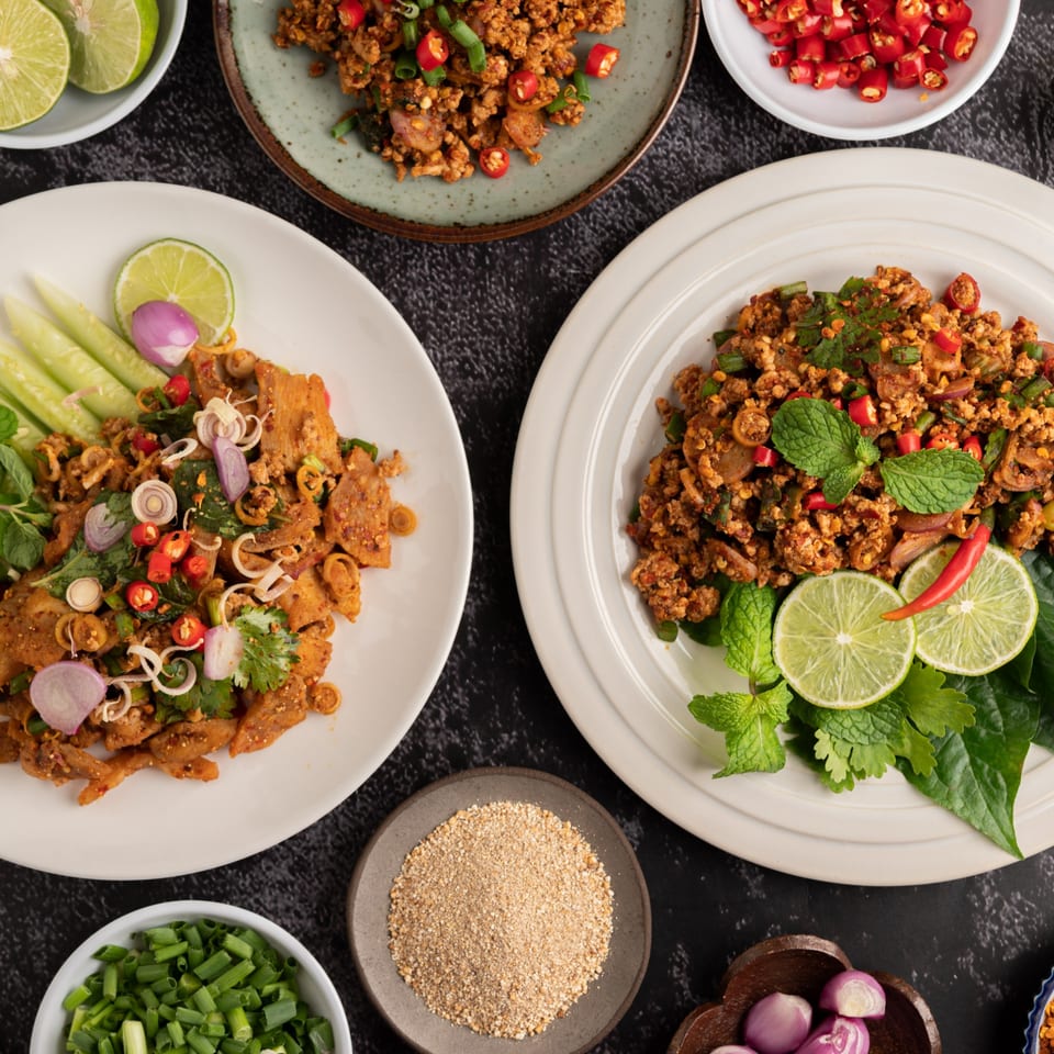 Two plates of Thai larb, one with pork and one with chicken, garnished with fresh herbs and lime.