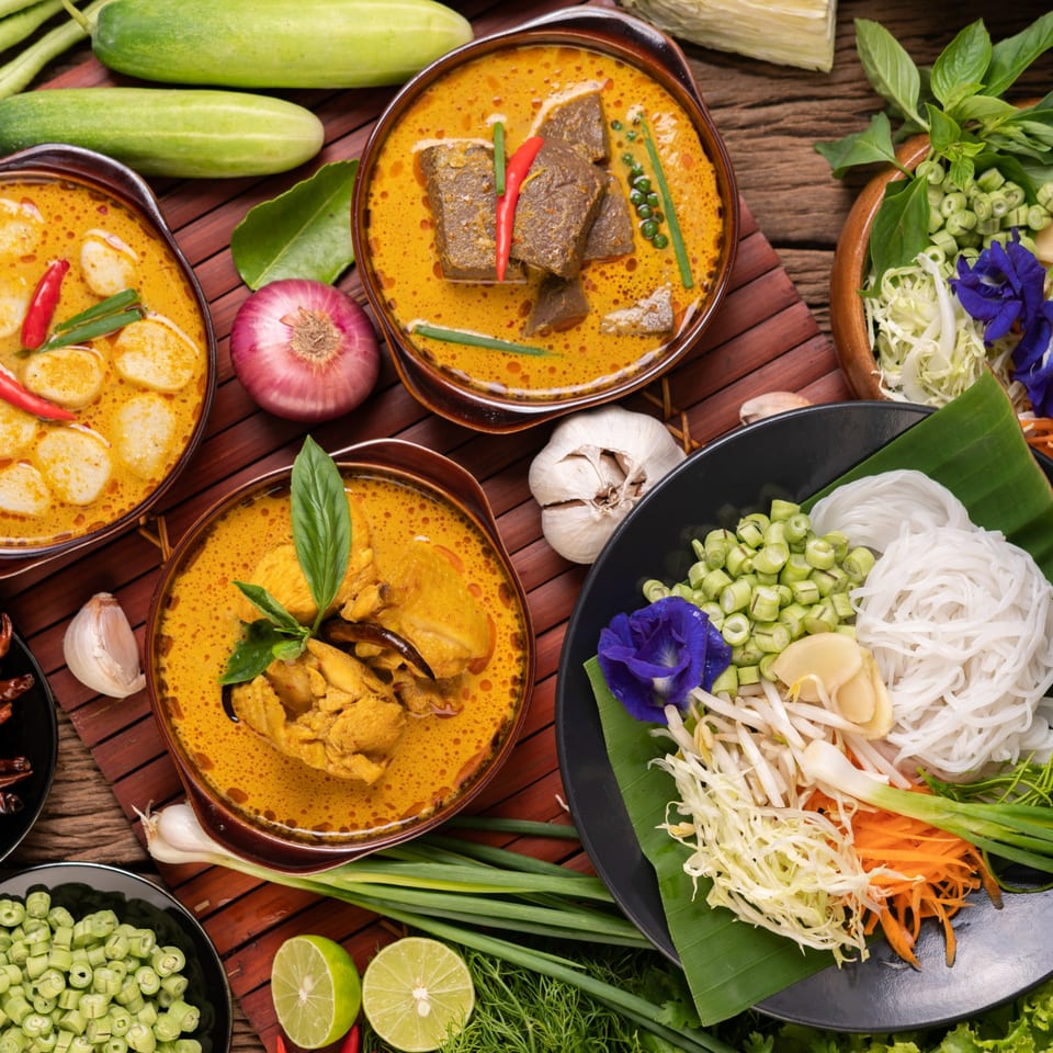 Three bowls of vibrant Thai curries, fresh vegetables, and rice noodles on a wooden table.